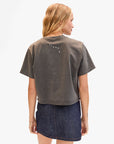 A woman with blonde hair wears the Clare Vivier Le Carre Tee, a loose, faded dark gray 100% cotton shirt with white text on the upper back, paired with a dark denim skirt. She stands with her back to the camera against a white background.