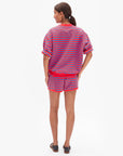 A woman with brown hair tied back wears the Clare Vivier Le Jogging Short—a red and blue striped cotton sweat short with an elastic waistband—paired with a matching short-sleeve sweatshirt and black sandals, set against a plain white background.
