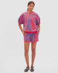 A woman stands against a plain background wearing Clare Vivier's Le Jogging Short and matching red and blue striped top with an elastic waistband, black sandals, a white necklace, and her hands in the shorts' side seam pockets.