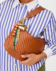 A person holds a Clare Vivier brown woven handbag with a wide strap, decorated with the Le Long Beaded Tassel and a vintage brass heart keychain, while wearing a blue and white striped shirt.