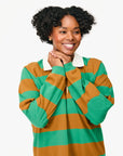 A cheerful person with curly hair wears the Le Rugby Dress by Clare Vivier, a green and brown striped heavyweight cotton piece featuring a crisp white collar. They rest their hands under their chin against a plain white background.