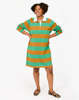A person beams, hands on hips, in Clare Vivier's Le Rugby Dress. This oversized, mustard and green 100% cotton dress features elbow patches for added style. They pair it with leopard-print flat shoes against a plain white backdrop.