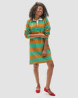 A smiling person with curly hair wears Clare Vivier's Le Rugby Dress, a striped green and orange piece crafted from 100% heavyweight cotton. Red flats complete the look against a plain white background.