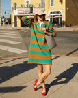 A woman in a Le Rugby Dress by Clare Vivier, featuring green and orange stripes with elbow patches, stands on a sunny sidewalk. Her beige cap and red shoes complement her checkered bag and beads, all set against a theater marquee that echoes her unique style.