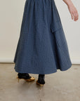 Wearing the Aquarius Cocktail Le Cargo Skirt—a long blue plaid cargo skirt with an elastic waist and pocket—this person pairs it with black patterned socks and gold heeled shoes while standing on a concrete floor.