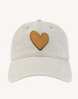 The Kerri Rosenthal KR Leather Heart Hat is a light beige cotton-twill adjustable hat with a brown heart-shaped patch on the front center, shown against a plain, light-colored background.