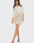 A woman poses against a white background wearing sunglasses, a long-sleeve white crop top, black sandals, and the Le Superbe Gypset Crochet Mirrorwed Mini Skirt.