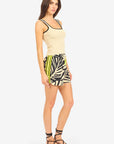 A woman with long brown hair stands sideways in a beige tank top and Le Superbe's BoHo Rhapsody Beach Boxer—high-waisted black and white shorts with yellow details, bold floral print—paired with strappy black sandals.