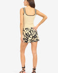 A woman stands with her back to the camera in a beige tank top with black trim and Le Superbe’s BoHo Rhapsody Beach Boxer, featuring an oversized floral motif and lightweight fabric, paired with black strappy sandals.