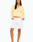 A woman poses against a plain background, wearing the Xirena Lillian Cardigan in pale yellow wool cashmere, white Bermuda shorts, a brown belt, brown boat shoes, and layered necklaces.