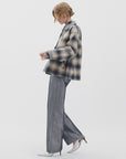A person with short wavy hair stands sideways against a white background in an oversized plaid jacket, Rag & Bone logan wide -metallic indigo jeans, and pointed white heels.