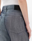 A person wears Rag & Bone's logan wide -metallic indigo jeans in lightweight, rigid denim with a faded gray-blue wash and a white ribbed tank top. The image highlights the jeans’ back pocket with a black waistband label and white stitching.
