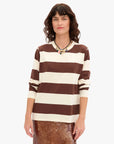 A woman with wavy brown hair wears the Clare Vivier LS Rugby Stripe Tee in brown and white, paired with a multi-colored necklace and shiny bronze skirt, standing against a plain white background.