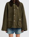 A woman wears the Denimist O/S Field Jacket in sturdy cotton—olive green with a dark brown corduroy collar, large front pockets, and silver snaps—paired with denim shorts. Her face is partially out of frame against a plain background.