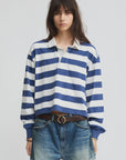 A young woman with long brown hair wears the Denimist Rugby LS Tee in Blue/Ecru Stripe, styled with denim shorts and a brown belt, standing against a plain light background.