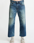 A person models Denimist’s HAISLEY SLOUCH BF JEAN—premium blue, mid-rise, straight-leg denim with light distressing—paired with white sneakers, standing against a plain white background. Upper body not visible.