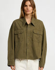A woman with long, light brown hair wears the Denimist CROPPED UTILITY SHIRT OLIVE, a boxy button-up with flap chest pockets, paired with white pants against a plain light background.