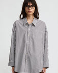 A person with long dark hair and glasses wears a relaxed fit Button Front Shirt by Denimist in black-and-white vertical stripes and brown pants, standing against a plain light gray background.