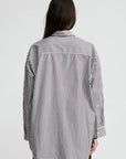 A person with long dark hair, seen from the back, wears Denimist’s Button Front Shirt in black-and-white vertical stripes and brown pants, both made from 100% cotton, set against a plain light background.
