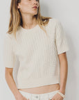 A woman with long, straight hair models the Denimist SHORT SLEEVE CABLE CREWNECK—an off-white, 100% cotton sweater—paired with white pants. She stands against a plain light background, one hand in her pocket.