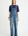 A person with long hair wears a navy tee and Denimist SLOUCH BIB OVERALL in Esher blue featuring a utility pocket, standing forward against a plain white background with a neutral expression and sneakers.