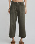 A person wearing Denimist’s Georgia Work Pant in olive green, featuring wide legs, 100% cotton fabric, and large front pockets, styled with a white cropped top and brown moccasin-style shoes against a plain light background.