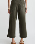 A person faces away, wearing Denimist's olive green 100% cotton Georgia Work Pant with a white top, brown moccasins, and bracelets on both wrists, set against a plain light background.