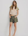 A woman stands against a white background wearing a color-blocked beige and green top, Denimist HAISLEY SHORT relaxed-fit women's cotton shorts in olive green, and white sandals, hands in her pockets with a relaxed expression.