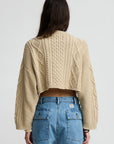 A person with long brown hair is seen from the back, wearing the Denimist Cropped Cable Sweater in beige and high-waisted blue denim shorts, standing against a plain light background.