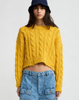 A woman stands against a plain light background wearing Denimist's Boxy Cable Sweater in yellow, paired with a navy bucket hat and high-waisted blue denim shorts with front pockets.
