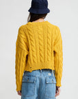 A person wearing the Denimist Boxy Cable Sweater in yellow, a navy bucket hat, and a denim skirt with large back pockets stands facing away against a plain white background.