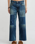 Someone stands against a plain light background wearing Denimist’s Lucy Boyfriend Jean—relaxed fit jeans with large, darker denim patches—paired with a purple tank top, moccasin-style shoes, and bracelets.