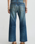 A person stands facing away, wearing Denimist's Lucy Boyfriend Jean with back pockets and a brown leather patch, a blue top, and brown platform sandals. Their hands rest by their sides, with bracelets on both wrists.
