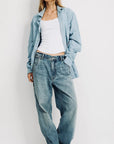 A woman stands against a plain background wearing the Denimist TERI WIDE LEG CARPENTER PANT, paired with a light blue denim shirt and white tank top. Her hands rest in her pockets, hair loose, highlighting relaxed style and soft cotton comfort.