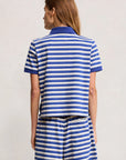 A person faces away, wearing Denimist's SHRUNKEN POLO—a blue and white striped cotton polo t-shirt and matching shrunken-fit shorts—against a plain light background.