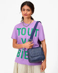 A woman in a lavender T-shirt, cream pleated skirt, and beaded necklace holds a blue bag with the Clare Vivier Adjustable Crossbody Strap, featuring cotton webbing, standing against a white background.