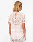 A woman with blonde hair is seen from behind wearing the Clare Vivier Lou Lou Top, a white, short-sleeve cotton lace blouse featuring a keyhole opening, neck tie closure, and sheer floral lace, paired with a matching skirt.