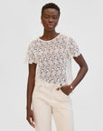 A woman with short hair stands against a plain light background, wearing the Clare Vivier Lou Lou Top—a white cotton lace short-sleeved blouse with a delicate neck tie—paired with light pants, gold earrings, and a bracelet.