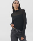 A woman with long brown hair wears the Rag & Bone luca dbl layer mix media mockneck ls top and faded black jeans, standing against a plain white background.