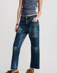 A person stands against a plain light background, hands in pockets, wearing a fitted gray tank top, Denimist Lucy Boyfriend Jeans with patchwork and frayed hems, and brown moccasin-style shoes.