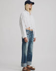 A person stands against a plain background wearing a navy baseball cap, a loose white cropped long-sleeve shirt, Denimist Lucy Boyfriend Jeans in 100% cotton with cuffs, and brown boat shoes showing part of the midriff.