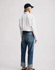 A person stands facing away, wearing Denimist's Lucy Boyfriend Jean with cuffs, a navy cap, white jacket, and brown sandals against a plain light background.