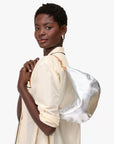 A woman in a light-colored shirt stands with her back slightly turned, looking at the camera and holding the Clare Vivier Lune, a metallic silver chrome-tanned leather shoulder bag with a brown strap.