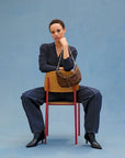 A person sits backwards on a wooden chair with red legs, wearing dark long sleeves, jeans, and black boots. They hold the Clare Vivier F25- Lune hair-on leather handbag with a knotted shoulder strap against a blue backdrop.