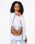 A person with curly hair holds Clare Vivier's Shortie Strap handbag, featuring a vintage brass finish and gold & silver studs. They wear a light blue shirt with white cuffs and light-colored jeans, looking to the side against a plain white background.