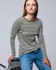 A woman with straight brown hair wears the Majestic Filatures Organic Cotton Stripe L/S Crewneck in green and white stripes with blue jeans, seated on a black chair against a light grey background.