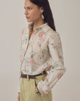 A woman with long dark hair wears the Narciso Shirt by Maison Hotel, an oversized cotton floral print top tucked into high-waisted khaki pants with a brown belt, earrings, and a patterned scarf. She stands in profile against a plain background.