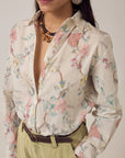 A woman wears the Narciso Shirt by Maison Hotel, a floral print top tucked into high-waisted beige pants with a dark belt. She accessorizes with a necklace and large round earring; only her head and upper body are visible.