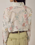 A person faces away, wearing the oversized Narciso Shirt by Maison Hotel—cotton with floral print—tucked into high-waisted pale green pants and a brown belt. Their dark hair is loose, accessorized with a large hoop earring.