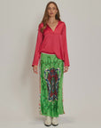 A woman with long brown hair wears a bright pink long-sleeved blouse, white boots, and the Me369 Blakely Slip Skirt—a green maxi skirt featuring a bold abstract pattern in red, blue, and black.